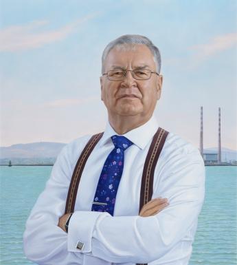 Three quarter length portrait of a man. He is wearing a white shirt, braces, and a blue tie. In the background we can see the sea, the mountains, and the Poolbeg towers.
