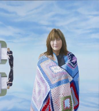 In the right foreground stands a female figure, wrapped in a colourful crochet blanket. In the background is a caravan, which is not hitched to anything. Leaning against it is a figure dressed in dark overalls. There is a plant beside the caravan's door, and the caravan, the figure and the plant are all reflected in the blue surface below.