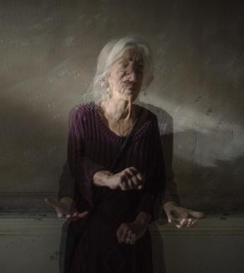 This portrait has the effect of a moving image within a still image. The subject is sitting in a shaft of light in a dimly lit background. She appears very animated, with her hands and head in motion. 