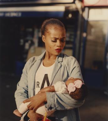 A woman dressed in a denim blazer and white t-shirt gazes down at the infant she cradles in her arms. The infant appears to be asleep, and has a large pink bow on her head. In the hand that is not holding the baby, the woman holds a red iced drink with a straw.