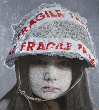 A child stares directly at the viewer. Her face is coated in a white dust-like substance, and she wears a helmet on her head, fastened under her chin. The helmet is covered in bubble wrap, and has red and white tape on it with the word 'Fragile'.