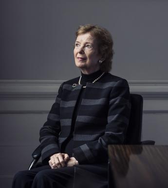 Former President of Ireland Mar Robinson sits beside a wooden table with her hands clasped in her lap. She is dressed in a grey and black striped jacket with a brooch at the lapel, and she gazes upwards and to her right.
