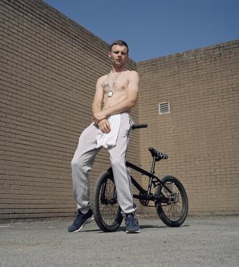 The subject of the portrait looks down a the camera lens, which is below him. He is leaning on the handlebars of a black bike which is behind him. He wears grey tracksuit pants and navy runners, and has a white tshirt tucked into his waistband. His hands are clasped in front of him, and he has a tatoo on his chest. Around his neck he wears a scapular and a rosary neads. Above his head, we see a section of blue sky above the brick wall behind him. 