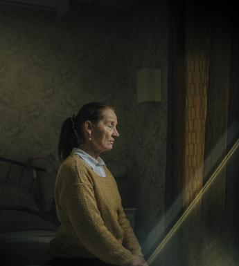 A woman in a mustard-coloured jumper over a white shirt sits on a bed with her eyes closed and her hands in her lap. She is turned towards a curtained window, and a shaft of light is coming into the darkened room in which she sits.