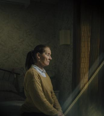 A woman in a mustard-coloured jumper over a white shirt sits on a bed with her eyes closed and her hands in her lap. She is turned towards a curtained window, and a shaft of light is coming into the darkened room in which she sits. 
