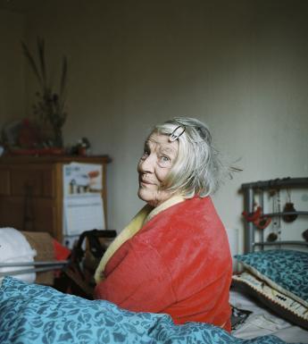 A woman in a red and yellow dressing gown sits on an unmade bed with a blue cover, looking at the viewer over her shoulder with an amused expression on her face. In the background we see a chest of drawers with a calendar attached to the side, and flowers and other belongings on top. The room is filled with signs of life - chairs, clothes, beads hanging on the bedspread etc.
