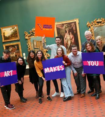 Group photo of men and women posing in an art gallery and holding colourful signs that say 'Apollo Project' and 'Art Makes You'.