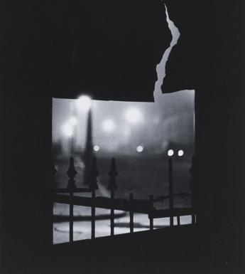 Black and white fine-art photograph of a gloomy city night scene viewed through a crack in a fence.