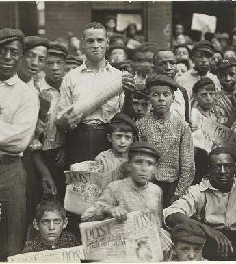 Black and white photo from 1908 showing a group of newsboys and men crowded together on the street
