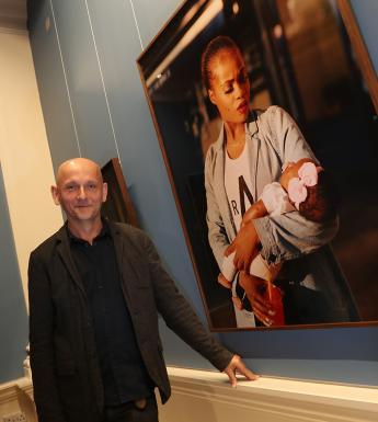 Photo of a man posing in a gallery beside a large fine-art photograph of a woman holding a child.