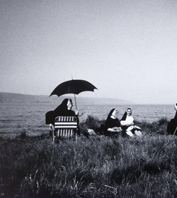 A group of nuns sit on a grassy patch overlooking the coast. Some sit on deckchairs, one with an umbrella over her head to shade her from the sun; others sit on blankets. 