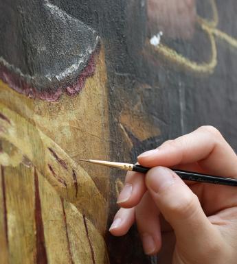 A conservator's hand holding a thin paintbrush in front of an oil painting