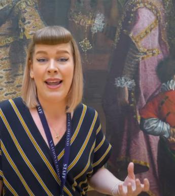 Film still of a blond woman wearing a striped dress speaking in front of a large oil painting