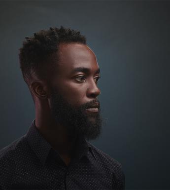A photographic portrait of a man. He is standing slightly turned away from the viewer, and dressed in a buttoned up shirt. He gazes to the right of the frame.
