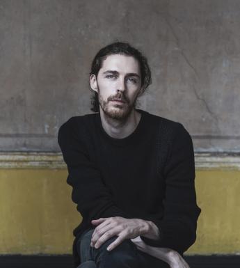 A photographic portrait. A man sits on a stool in front of a yellow and grey wall, with paint peeling. He wears a black round neck jumper, and dark jeans. His hands look as if they are in motion.