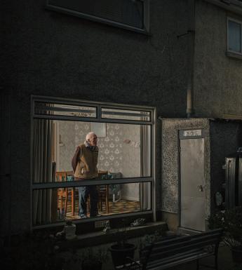 The viewer is looking in through the window of a house at night. A man stands in the illuminated window of a dining room, gazing out, hands behind his back.