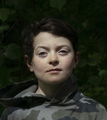 A photographic portrait of a boy. He looks directly at the camera, and is wearing a camouflage hoodie.