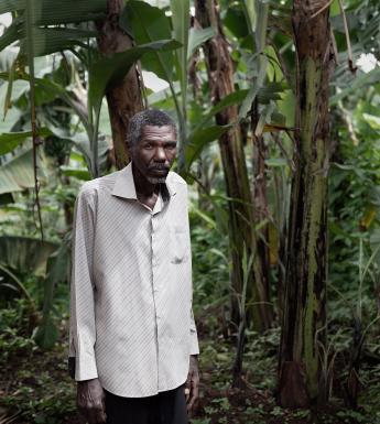 A man stands, with what thick foliage behind him. He wears a long sleeved buttoned shirt, untucked, and black pants. His arms are by his side, and he looks directly at the viewer.