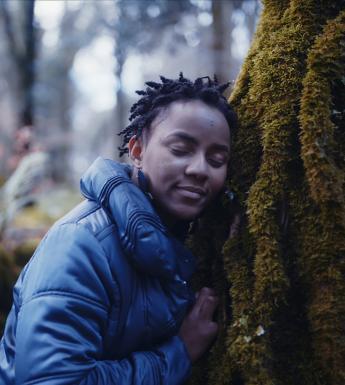 A still from a documentary film. A woman leans against a moss-covered tree, eyes closed and her ear pressed up against the trunk.