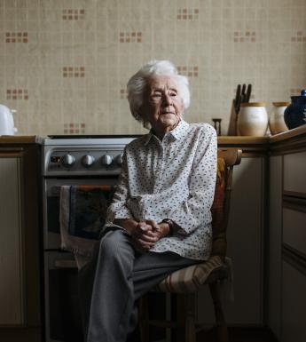 A woman sits on a chair in her kitchen, her hands clasped in her lap. Her legs are crossed, and she looks down, away from the viewer.  