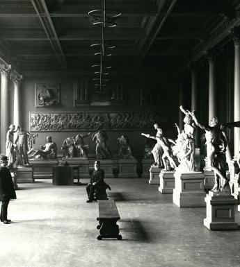 A black and white photograph of a room filled with plaster casts of sculptures. There are two me in the room - one sits on a bench in the middle of the room, the other stands on the left, facing the seated man.