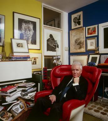 Photo of Alexander Walker in his apartment sitting in a red leather armchair with lots of art hanging on the walls