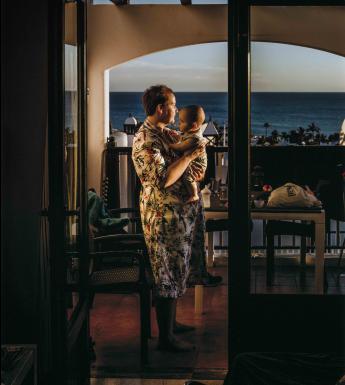 Portrait photograph of a woman wearing a robe and holding baby, standing in a room with a large window with a view of the sea