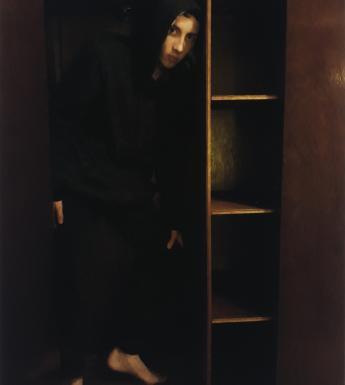 Portrait of a young man wearing black trousers and black top with hood up standing in the shadows in an empty wardrobe