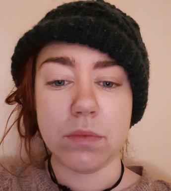Video still, close-up view of a woman's face. She is wearing a black woolen hat and her eyes are cast downwards.