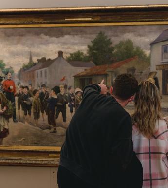 A photograph showing a man and woman standing looking at a large oil painting. Their heads are close together and the man is pointing at something in the painting.