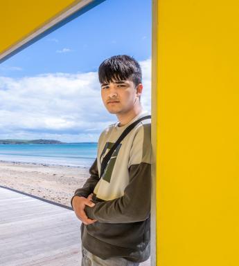 Photograph of a male figure with dark hair and a dark green jumper leaning against a yellow structure with his back to the beach