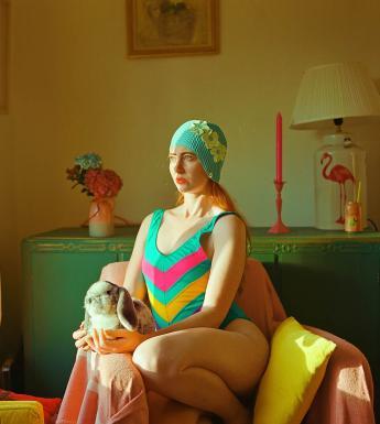 Photograph of a female figure with red hair wearing a teal striped swimsuit and hat sitting in a pink chair holding a toy rabbit