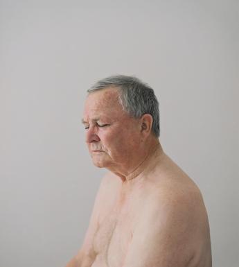 Photograph of an older man's head and shoulders with grey hair and his eyes closed sitting nude on a grey background