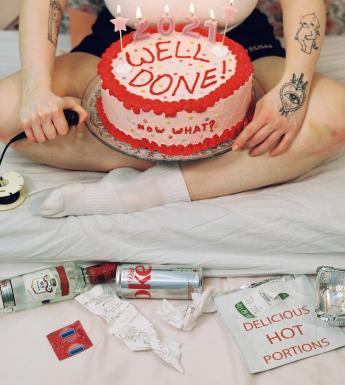 Photograph of a figure with tattoos sitting cross-legged on a bed holding a cake with various rubbish nearby