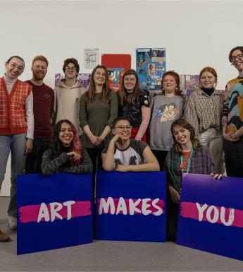 Photograph of a mixed group of young people, with three purple signs with the statement 'Art Makes You'