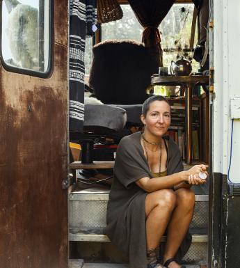 A photograph of a woman sitting on the steps of a caravan. She is wearing a long grey cardigan and has bare legs. Her hands are clasped in front of her.