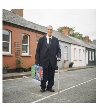 A man in a large, baggy black suit stands in front of a row of cottages. He is leaning on a crutch. In the other hand he carries a blue shopping bag. 