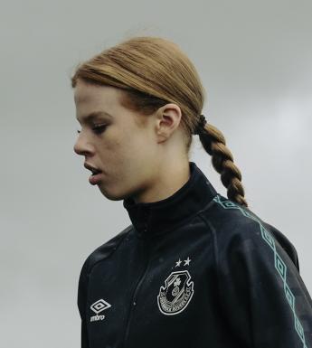 A young woman in a crested tracksuit top stands in profile, eyes cast down. Her red hair is tied back in a long plait.