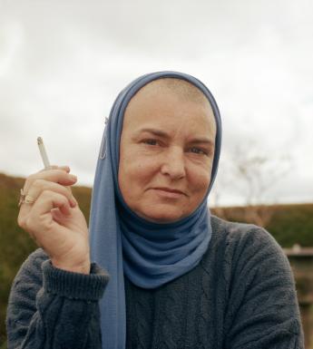 A photographic portrait of a a woman. She wears a dark blue knitted jumper and a blue headscarf. She holds a cigarette aloft in one hands. Behind, a grey sky and some green fields. 