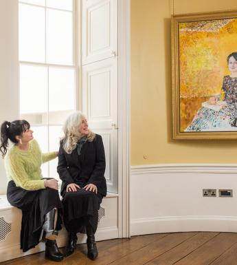Photograph of two women, one wearing black, the other wearing a green top, sitting in a yellow room looking at a painting