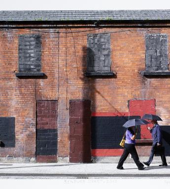 A photograph of a row of red brick houses with the windows and doors boarded up. Two people walk by in opposite directions, both holding umbrellas.
