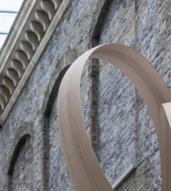 Cropped image of a curved grey wooden sculpture in front of a grey stone wall.