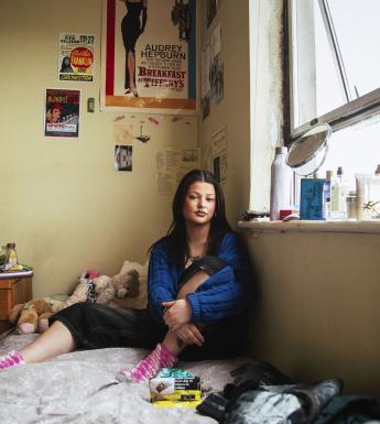 A photograph of a young woman sitting in her bedroom, surrounded by her belongings. On the wall, a number of posters and cuttings hang including a Breakfast at Tiffany's poster; on the bed itself are a few stuffed animals, and various cosmetics line the window sill.