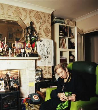 Shane MacGowan sits on a green armchair beside a fireplace. The mantle is filled with objects including a buddha, a picture of Jesus and other religious figures. A statue of Phil Lynott stands on the gas fire.