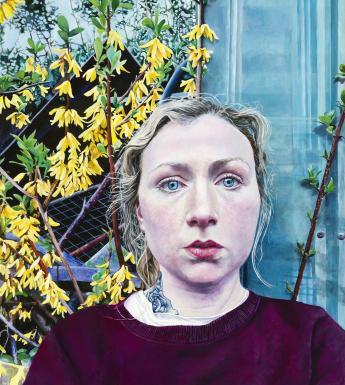 A woman with fair hair looks directly at the viewer. She wears a dark red top, with a white t-shirt underneath. A tattoo can be seen on her neck. Behind her the metal of a garden shed, and a bright yellow plant in bloom.