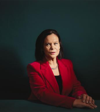 Dressed in a red jacket and seated at at table with her hands clasped in front of her, Mary Lou McDonald sits in front of a dark green wall.