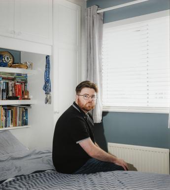 A young man sits on the bed in a neat bedroom, looking over his shoulder at the viewer. The shelf over the bed is filled with books and other belongings.