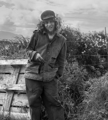 A black and white photograph of a man standing in a windswept hilly landscape, with a wooden palette directly behind him. He has long curly hair underneath a cap, and is laughing at the camera.
