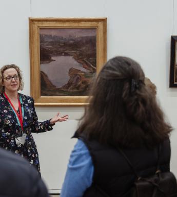 A guide stands in front of a painting, talking to a group of people who are looking at her. 