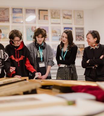 A group of young people standing around discussing prints and painting on a table.